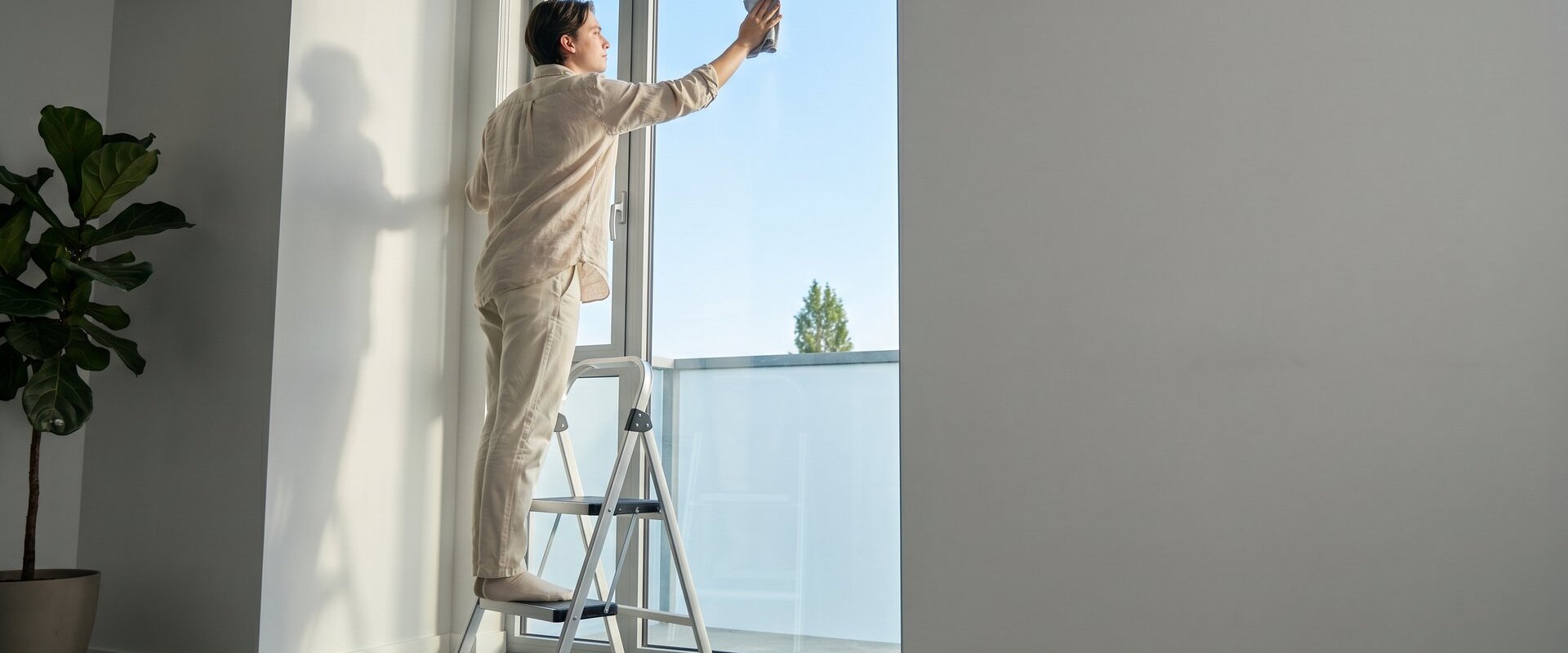 Person reinigt auf einer Trittleiter ein hohes Fenster in einer hellen Wohnung